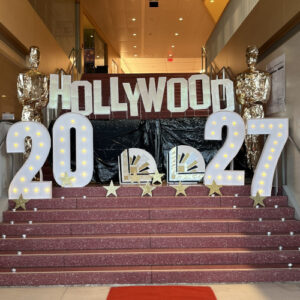 Staircase decorated for a Hollywood-themed junior class event, featuring large illuminated marquee numbers reading "2027," a "Hollywood" sign in the style of the iconic Los Angeles landmark, gold Oscar statuette replicas, and gold star decorations on the steps.