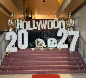 Decorative staircase display for the BHS Class of 2027 Junior Semi, featuring illuminated marquee numbers reading "2027," a hand-lettered "Hollywood" sign, gold star cutouts, and two gold Oscar-style statues flanking the steps. A red carpet is visible at the base of the stairs.